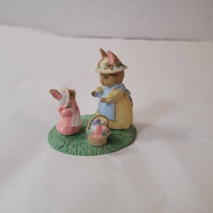 May include: A figurine set featuring two bunnies and a basket of eggs. One bunny wears a pink dress and bonnet, while the other wears a yellow dress, blue apron, and a hat. The figurines are on a green base.