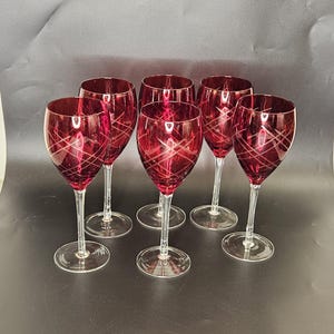 May include: Six ruby red wine glasses with clear stems and bases. Each glass features a crisscross etched design. The glasses are arranged on a dark surface, showcasing their elegant form and color.