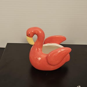 May include: A glossy, coral-pink ceramic planter shaped like a flamingo. The bird has a curved neck, a yellow beak with a small dark eye, and raised wing details on the sides. The interior of the planter is white, and the overall finish is smooth.