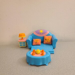 May include: A blue toy sofa with a yellow sun and pink flowers on the back, two orange pillows, and a blue table with a yellow teapot and four orange cups. A blue nightstand with a yellow lamp is on the left.