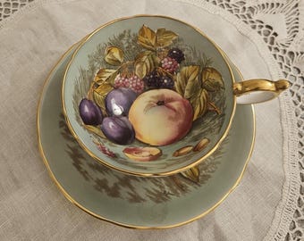 May include: A vintage teacup and saucer set with a light green background and gold trim. The teacup interior features a detailed illustration of various fruits, including plums, peaches, and blackberries. The handle is gold.