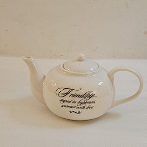 May include: A white ceramic teapot with a rounded body, a curved spout, and a loop handle. The teapot has a lid with a small knob. The words "Friendship... steeped in happiness, warmed with love" are written on the front.