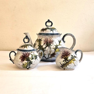 May include: A three-piece ceramic tea set featuring a teapot, sugar bowl, and creamer. The set is decorated with a thistle motif in shades of burgundy, green, and white, with blue trim. The teapot has a curved handle and a lid with a ring-shaped handle.