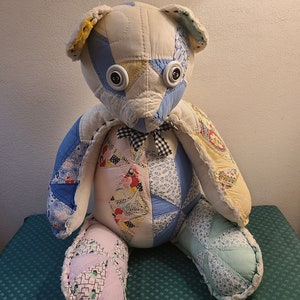 May include: A patchwork quilt teddy bear with blue, yellow, and pink fabric. The bear has large black button eyes and a black and white bow tie.