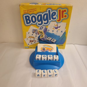 May include: Boggle Jr. game box with a blue and yellow design. The box shows a picture of a school bus and the words "Boggle Jr." and "Your preschooler's first Boggle game". The game includes a blue and yellow game board with letter tiles and a card with the word "DUCK" on it.