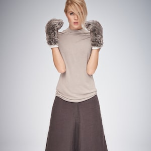 May include: A woman wearing a beige turtleneck top, wide-leg brown pants, and furry gray gloves with white cuffs. The outfit is simple and modern, suitable for fall or winter wear.
