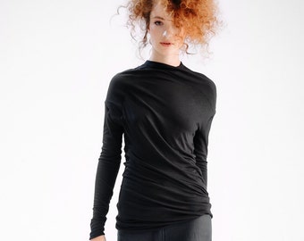 May include: A woman with curly red hair is wearing a black long-sleeved shirt and black pants. The shirt has a mock neck and the pants are loose fitting.