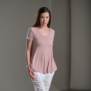 Puede incluir: Una mujer con una camisa rosa de manga corta y pantalones blancos. Lleva sandalias negras y tiene el pelo largo y casta&ntilde;o.