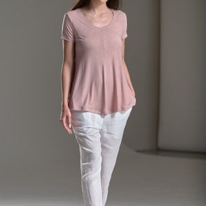 Puede incluir: Una mujer con una blusa de color rosa claro de manga corta, holgada, con cuello en V y pantalones de lino blancos. Lleva sandalias negras.
