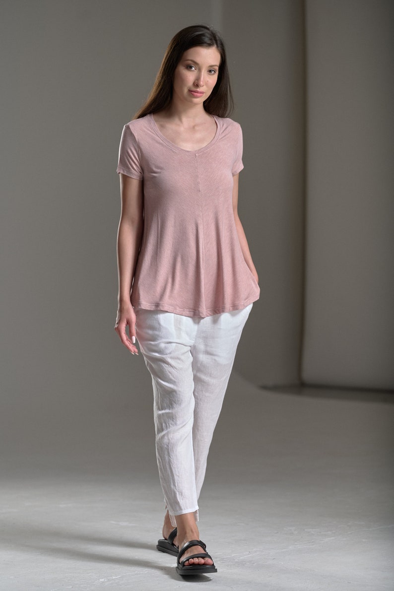 Puede incluir: Una mujer con una blusa de color rosa claro de manga corta, holgada, con cuello en V y pantalones de lino blancos. Lleva sandalias negras.