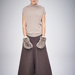 May include: A woman wearing a beige short-sleeved top, brown wide-leg pants, and faux fur mittens. The outfit is paired with sandals. The background is a solid light gray.
