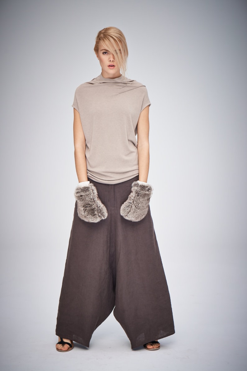 May include: A woman wearing a beige short-sleeved top, brown wide-leg pants, and faux fur mittens. The outfit is paired with sandals. The background is a solid light gray.