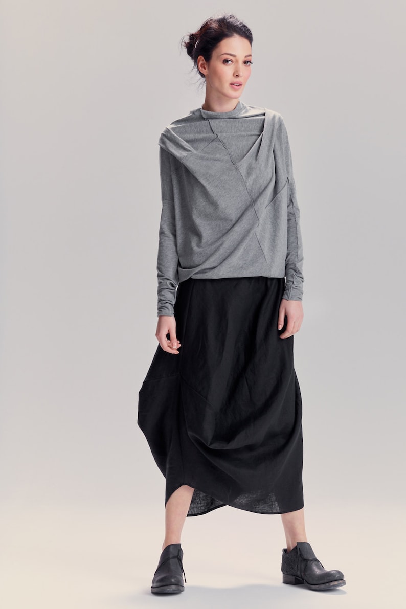 May include: A woman wearing a gray long-sleeved shirt with a draped neckline and a black, flowing skirt. She is wearing black ankle boots.