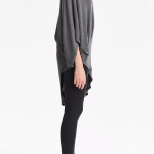 K&ouml;nnte beinhalten: Ein graues, lang&auml;rmeliges Tunika-Oberteil mit drapiertem Design und einem hohen Ausschnitt. Das Oberteil wird mit schwarzen Leggings kombiniert.