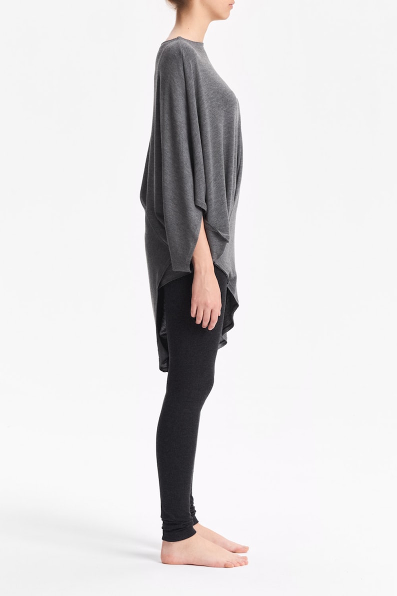 K&ouml;nnte beinhalten: Ein graues, lang&auml;rmeliges Tunika-Oberteil mit drapiertem Design und einem hohen Ausschnitt. Das Oberteil wird mit schwarzen Leggings kombiniert.