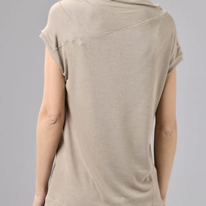 May include: A beige short-sleeved t-shirt with a unique neckline and a diagonal seam across the upper back. The shirt is made of a soft, neutral-toned fabric. A brown hat is visible in the image.