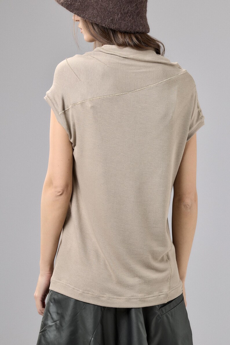 May include: A beige short-sleeved t-shirt with a unique neckline and a diagonal seam across the upper back. The shirt is made of a soft, neutral-toned fabric. A brown hat is visible in the image.