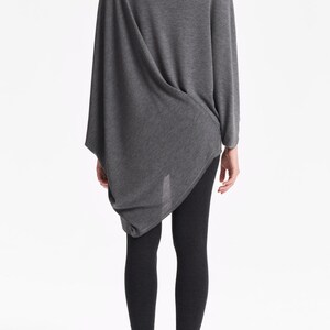 K&ouml;nnte beinhalten: Ein graues, lang&auml;rmliges Tunika-Oberteil mit drapiertem Design und lockerer Passform. Das Oberteil wird mit schwarzen Leggings kombiniert.