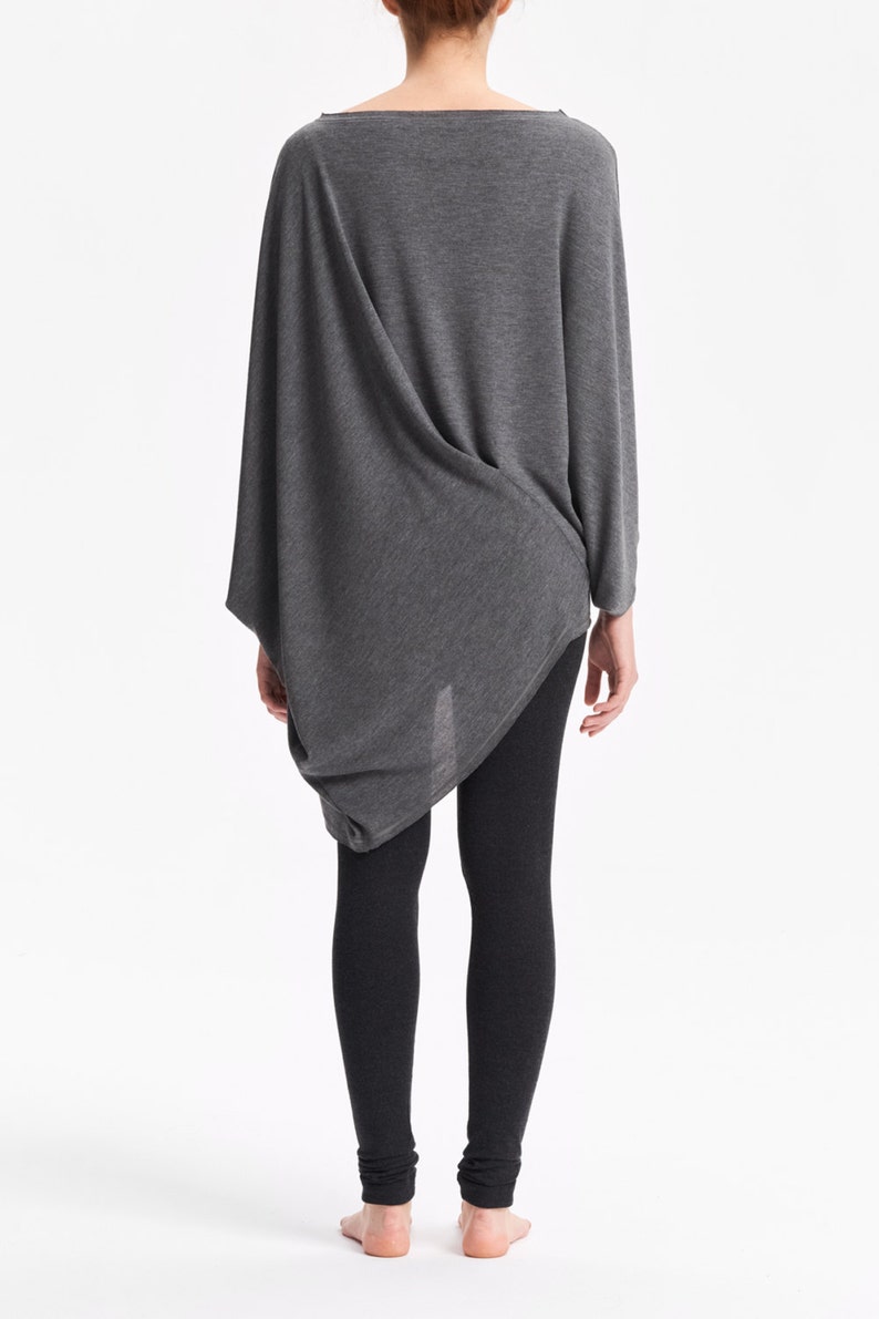 K&ouml;nnte beinhalten: Ein graues, lang&auml;rmliges Tunika-Oberteil mit drapiertem Design und lockerer Passform. Das Oberteil wird mit schwarzen Leggings kombiniert.