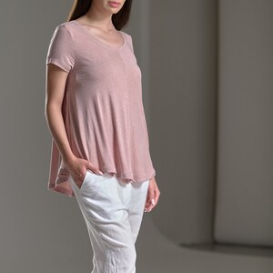 Puede incluir: Una mujer con una camiseta de manga corta de color rosa claro y holgada y pantalones de lino blancos.