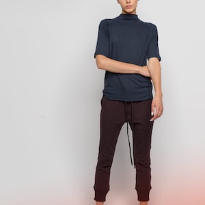 May include: A woman wearing a dark blue mock neck shirt with short sleeves and dark brown pants. She is wearing black sandals and has her hair pulled back.