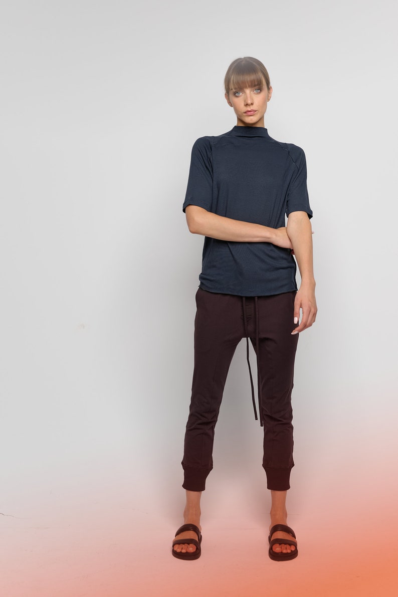 May include: A woman wearing a dark blue mock neck shirt with short sleeves and dark brown pants. She is wearing black sandals and has her hair pulled back.