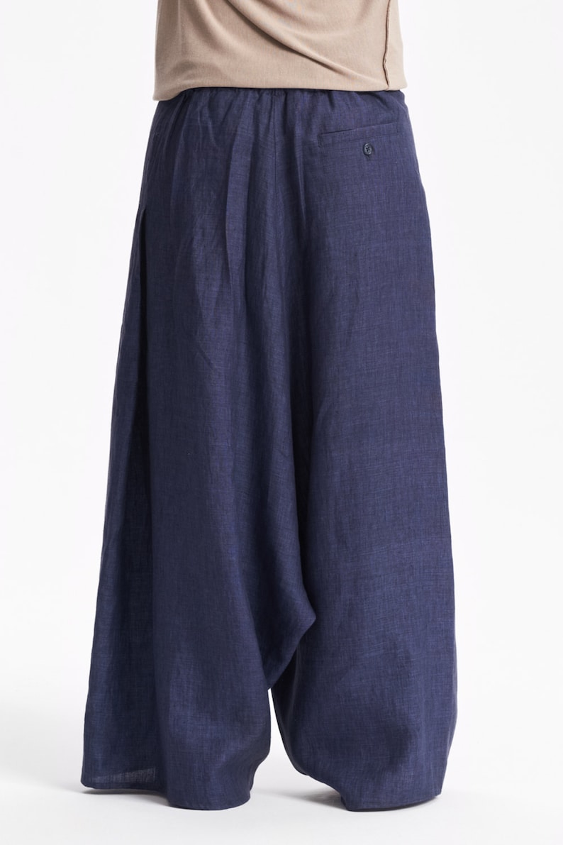 May include: Wide-leg navy blue trousers with a loose, flowing silhouette and a gathered elastic waistband. The pants feature a visible single button detail on the upper right side and a textured fabric finish, paired with a plain beige short-sleeve top.