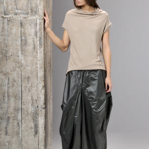 May include: A woman's fashion ensemble featuring a brown felt hat, a beige short-sleeved top, and a dark gray, voluminous skirt. The outfit is paired with dark shoes. The model is standing next to a concrete pillar.