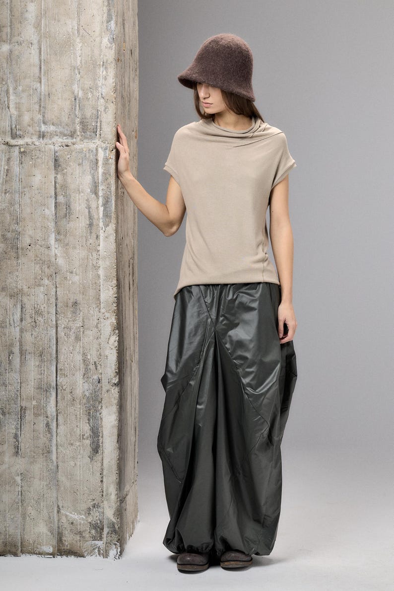 May include: A woman's fashion ensemble featuring a brown felt hat, a beige short-sleeved top, and a dark gray, voluminous skirt. The outfit is paired with dark shoes. The model is standing next to a concrete pillar.