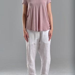 Puede incluir: Una mujer lleva una camiseta rosa de manga corta y pantalones de lino blancos. Lleva sandalias negras.