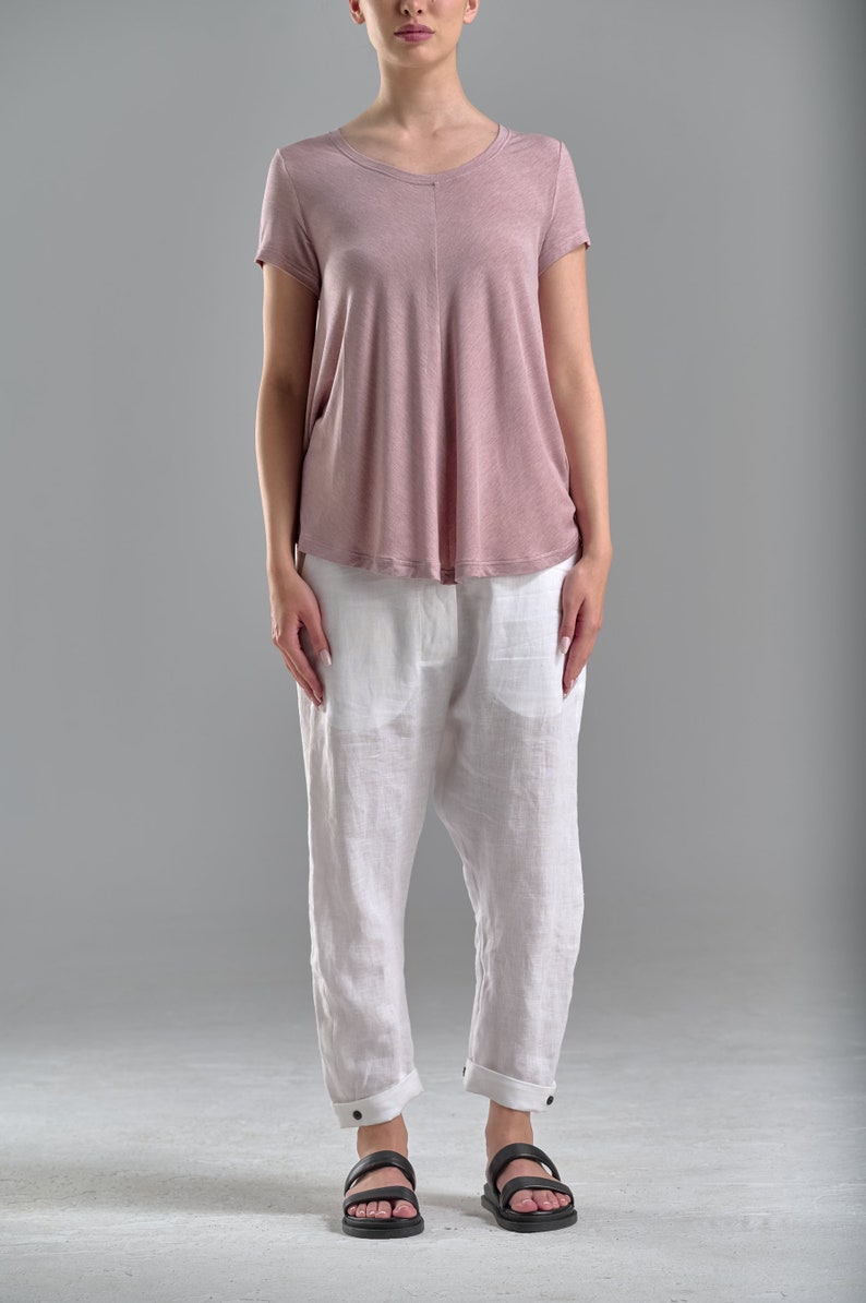 Puede incluir: Una mujer lleva una camiseta rosa de manga corta y pantalones de lino blancos. Lleva sandalias negras.