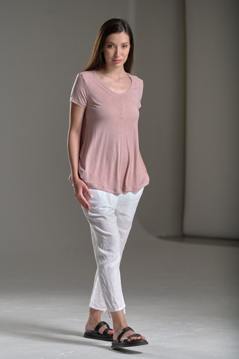 Puede incluir: Una mujer con una camisa rosa de manga corta y pantalones blancos. Lleva sandalias negras y tiene el pelo largo y casta&ntilde;o.