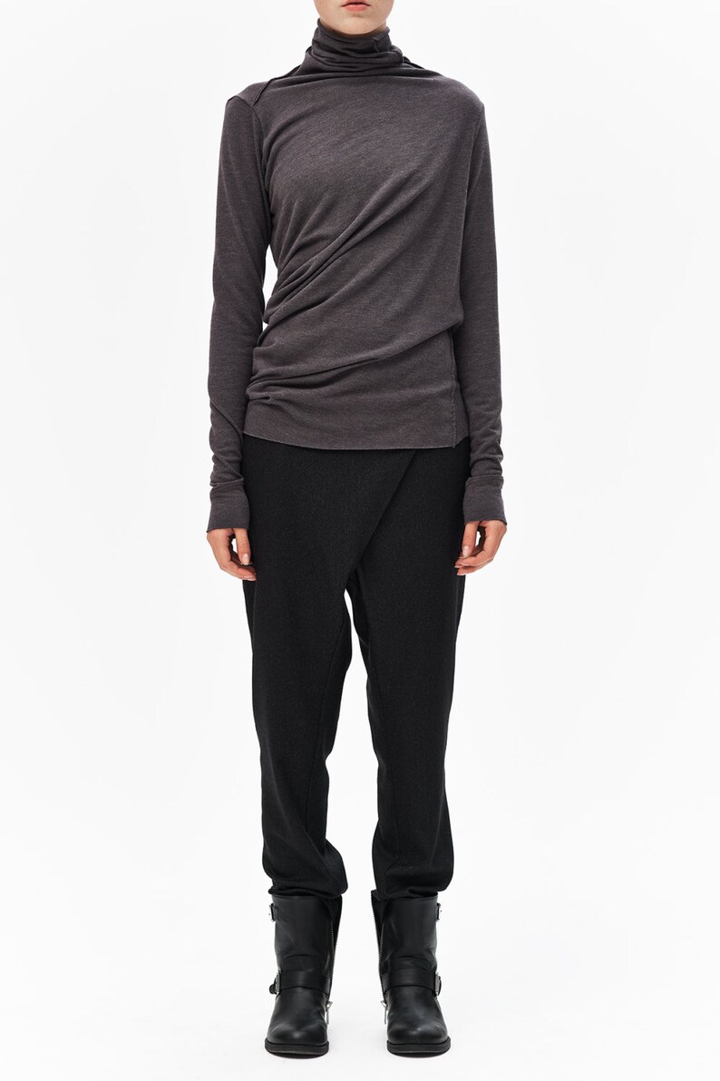 Puede incluir: Una camisa gris de cuello alto de manga larga con un dise&ntilde;o drapeado y pantalones negros. La persona lleva botas negras de tobillo.