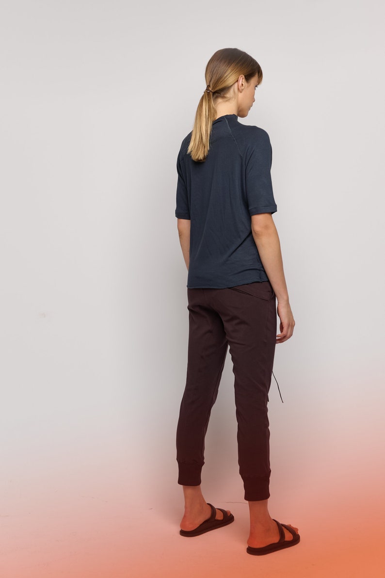 May include: A woman wearing a dark blue short-sleeved shirt and dark brown pants. She is standing with her back to the camera and her hair is pulled back in a ponytail.