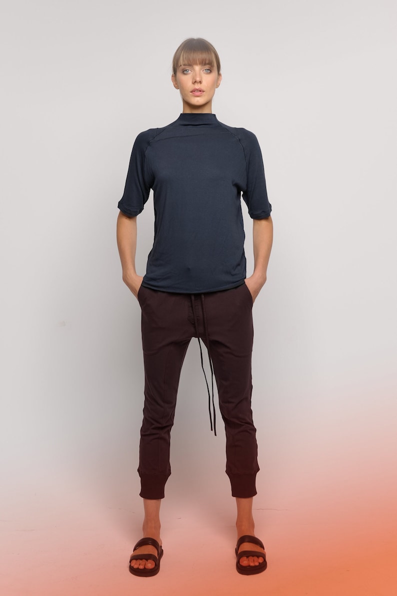 May include: A woman wearing a dark blue mock neck shirt with a relaxed fit and dark brown pants with a drawstring waist. She is wearing black sandals.