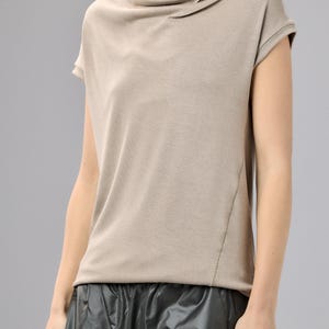 May include: A beige short-sleeved top with a draped cowl neckline. The top has a relaxed fit and a subtle seam detail on the side. The model is wearing a black skirt. This top is suitable for casual wear.