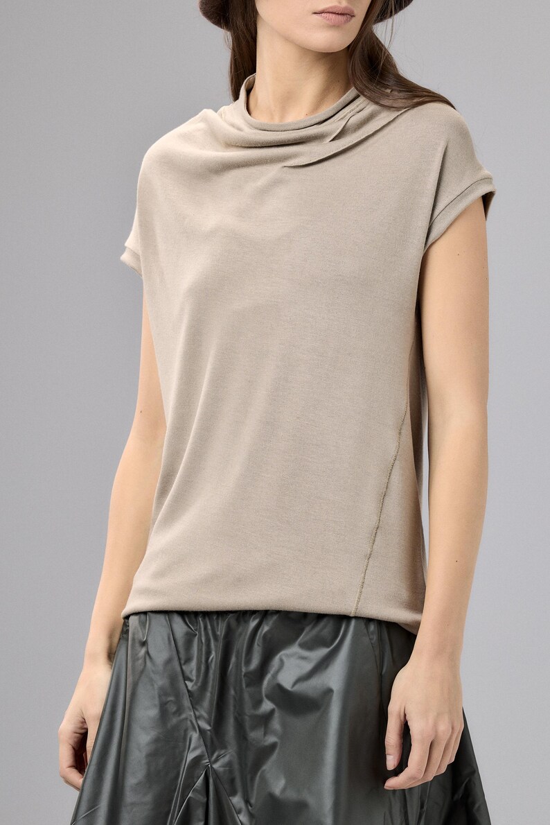 May include: A beige short-sleeved top with a draped cowl neckline. The top has a relaxed fit and a subtle seam detail on the side. The model is wearing a black skirt. This top is suitable for casual wear.