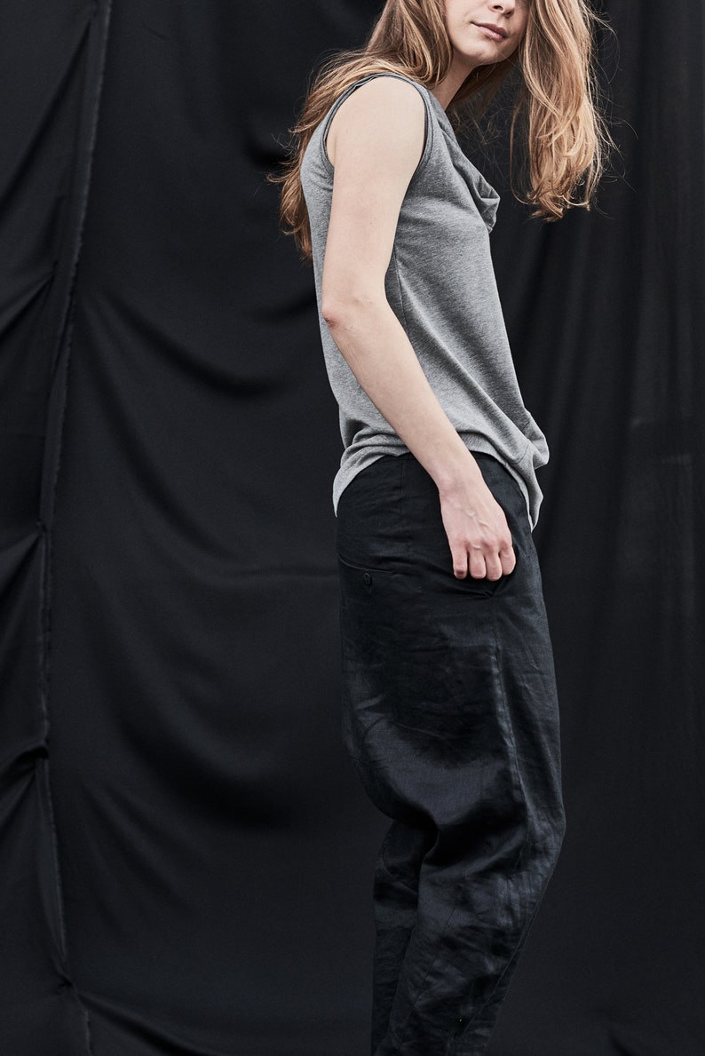 May include: A woman wearing a gray sleeveless top and black pants. The top has a cowl neck and the pants are loose fitting.