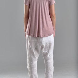 Puede incluir: Una mujer lleva una camiseta de color rosa polvoriento de corte holgado con mangas cortas y pantalones de lino blancos. Lleva sandalias negras.