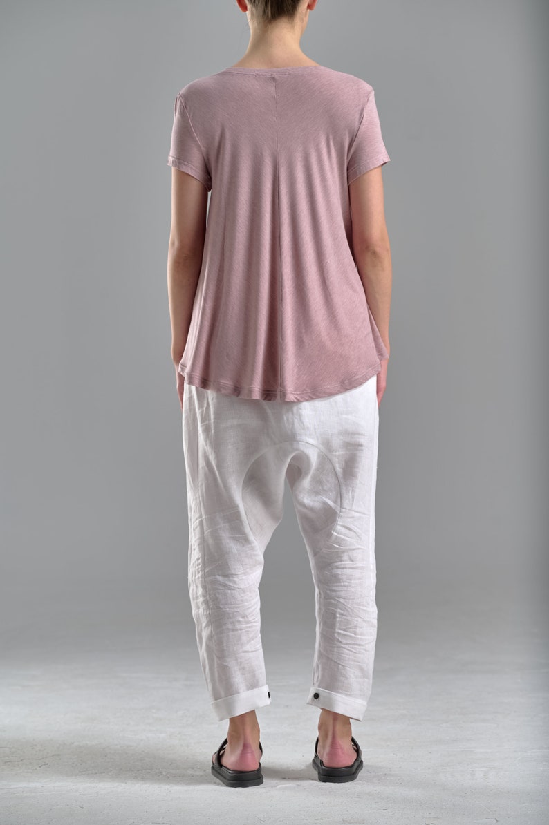 Puede incluir: Una mujer lleva una camiseta de color rosa polvoriento de corte holgado con mangas cortas y pantalones de lino blancos. Lleva sandalias negras.