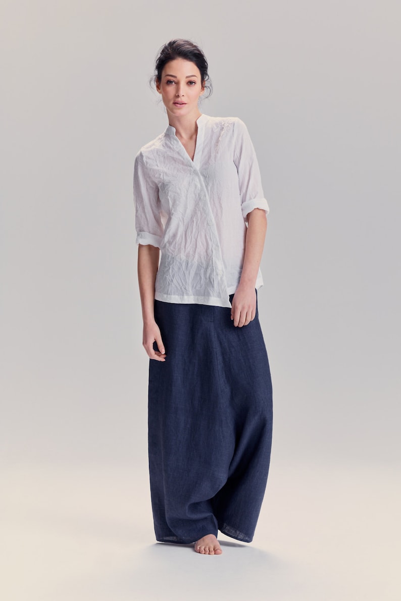 May include: A white long-sleeved shirt with a textured, crinkled finish and a mandarin collar, paired with a pair of loose-fitting, floor-length dark navy blue trousers. The shirt features a V-neckline and sleeves that are rolled up to the elbows.