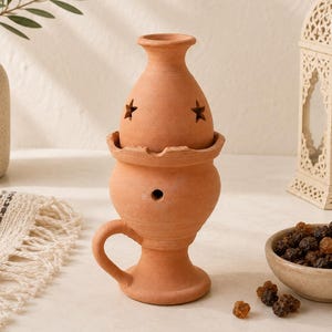 Può includere: Un bruciaincenso in terracotta dal design in due pezzi con coperchio rimovibile, intagli a forma di stella, un piccolo foro circolare laterale e un manico curvo. La base è a piedistallo, realizzata in argilla naturale opaca.