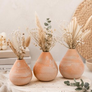 Op de afbeelding: Drie terracotta vazen met droogbloemarrangementen. De vazen hebben een druppelvorm en een getextureerde, lichtoranje afwerking. Elke vaas bevat een ander arrangement van gedroogde bloemen en grassen, wat een natuurlijke, bohemien uitstraling creëert.