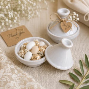 May include: Two white ceramic tajine-shaped containers, one open and filled with white and gold foil-wrapped sweets and small white flowers, the other closed with a twine-tied heart-shaped tag reading Thank You with a small heart symbol.
