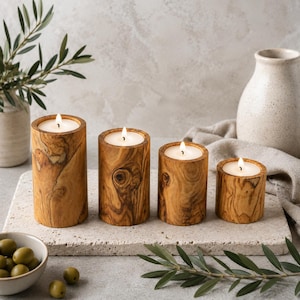 Può includere: Quattro portacandele in legno con candele bianche accese. I supporti sono cilindrici, realizzati in legno con un motivo a venature naturali. Le candele sono disposte in fila su una superficie di pietra, con un vaso e un ramo d'ulivo sullo sfondo.