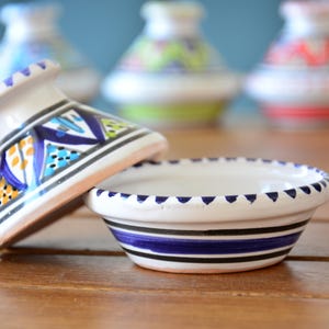 Może przedstawiać: Biała ceramiczna tagine z niebieskimi i zielonymi wzorami geometrycznymi. Tagine jest otwarta, a pokrywka spoczywa na białej ceramicznej misce z niebieskimi paskami.