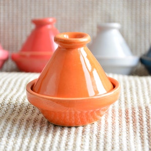 Könnte beinhalten: Ein Satz von fünf Keramik-Taginen in verschiedenen Farben, darunter Orange, Rosa, Weiß und Blau. Die Taginen sind in einer Reihe auf einer gewebten Oberfläche angeordnet.