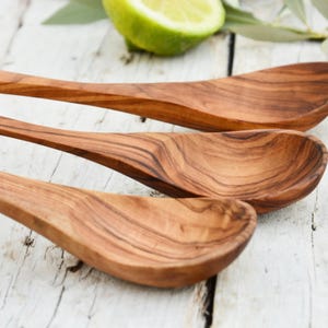 May include: Three wooden spoons with a natural wood grain pattern are arranged diagonally. The spoons are a warm brown color and appear to be made of olive wood. A lime half and green leaves are in the background.