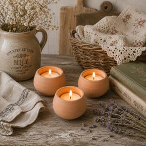 May include: Three small, round, terracotta-colored candle holders, each containing a lit white tea light candle. The holders have a smooth, matte finish and a simple, spherical shape, arranged on a wooden surface with other decorative items.
