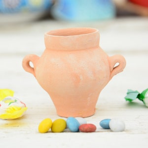 Pode incluir: Um pequeno pote de terracota com duas alças, sentado em uma superfície branca com doces coloridos espalhados ao redor.